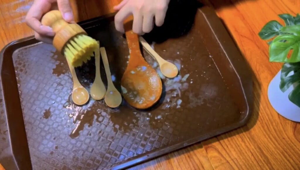 Hajar using a bamboo dish brush to clean wooden kitchen utensils on a baking tray